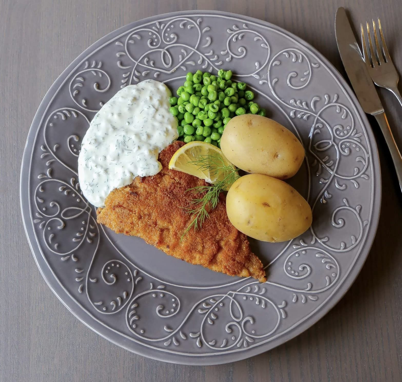 Breaded fish fillet on a grey patterned plate with dill sauce, green peas, boiled potatoes and a lemon wedge